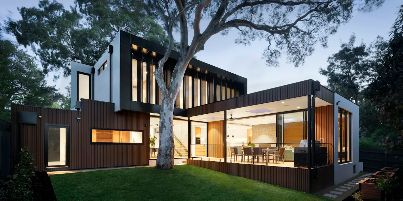 Modern house with lit windows under a large tree at dusk