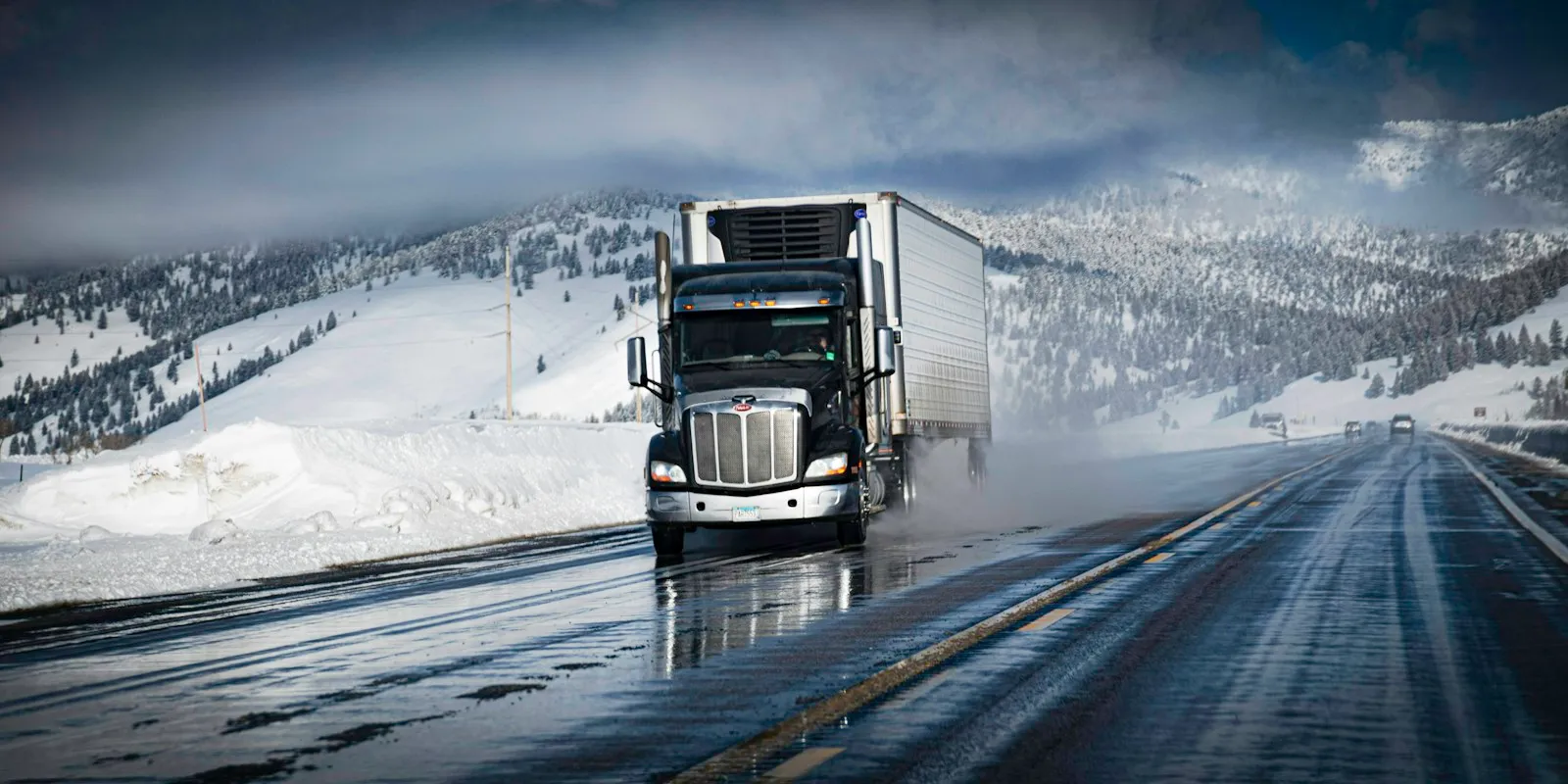 Refrigerated semi truck driving on a wet highway through snowy mountains
